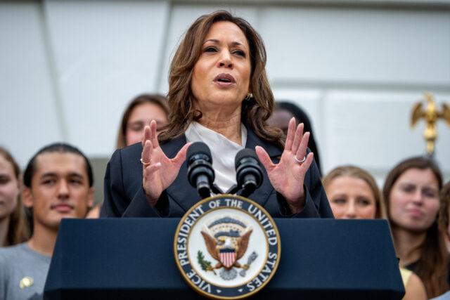 Kamala Harris, in a navy blue suit and white blouse, holds both hands in front of her, gesturing as she speaks at a podium, with a group of young people standing behind her.