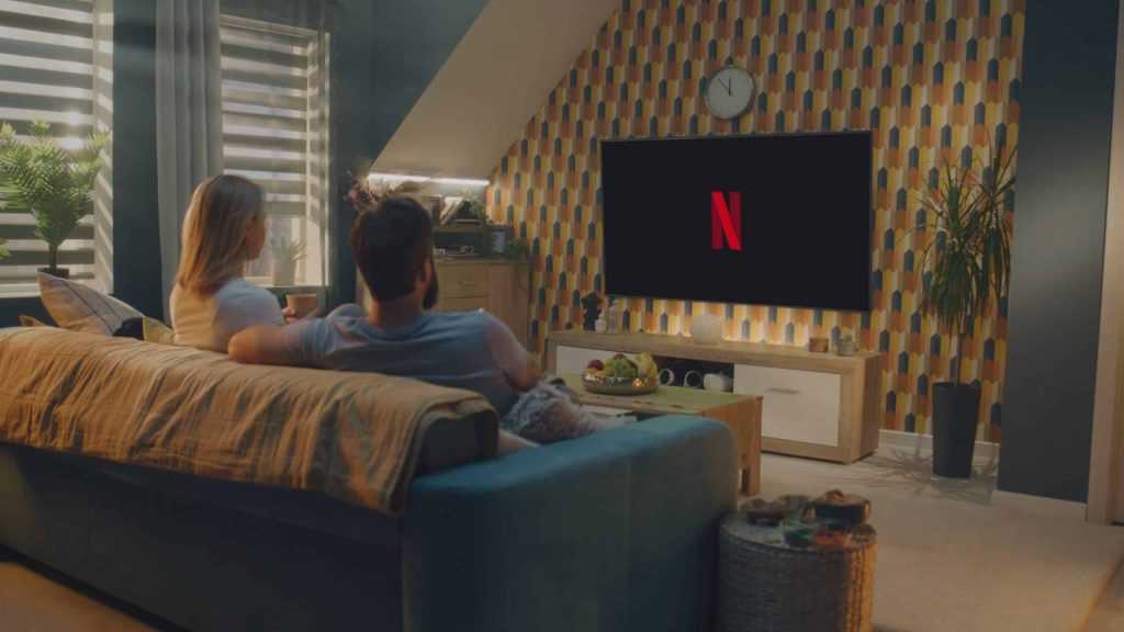 ZAPORIZHYA, UCRÂNIA - 7 DE JUNHO DE 2024: Casal bebe chá e começa a assistir filme ou programa de TV no serviço de streaming Netflix sentado no sofá em casa e descansando. Logotipo da Netflix exibido na grande tela da TV